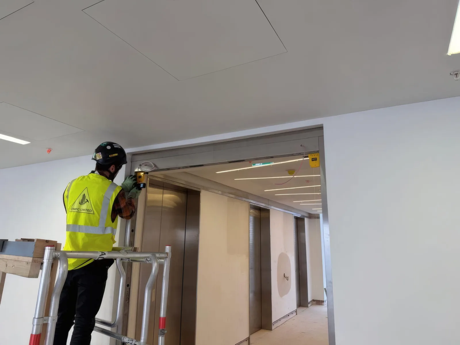 An OMFC International installer setting out above a door frame at the lift lobby, using a podium platform.