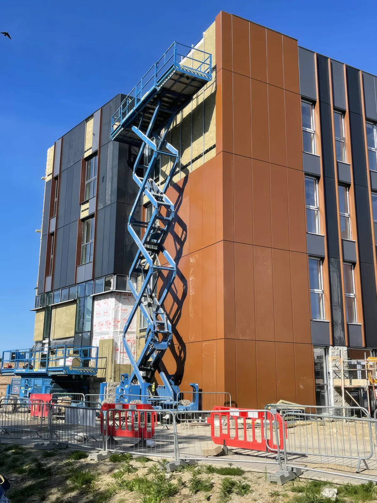 Copper-coloured cladding going up at a corner of the building, with a scissor lift in place.